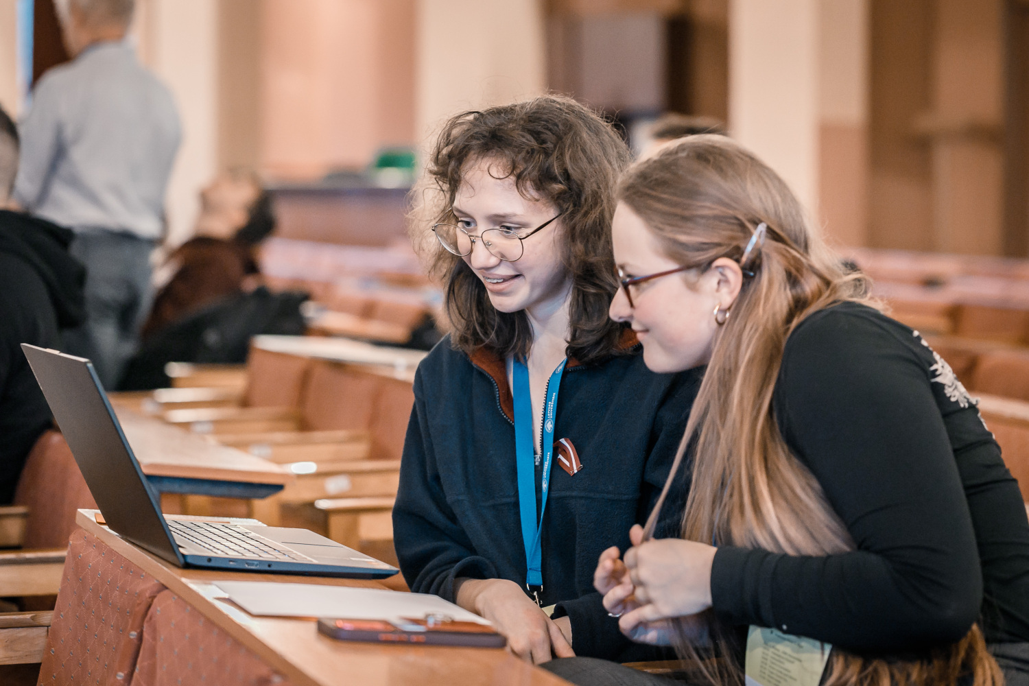 Two students are sitting together in a lecture hall, looking at a laptop